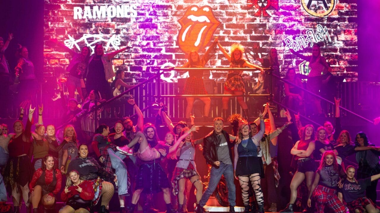 A large group of performers in punk and rock-inspired costumes pose energetically on stage, with colourful lighting and a brick wall backdrop featuring band logos such as AC/DC, The Ramones, The Rolling Stones, and We Will Rock You.