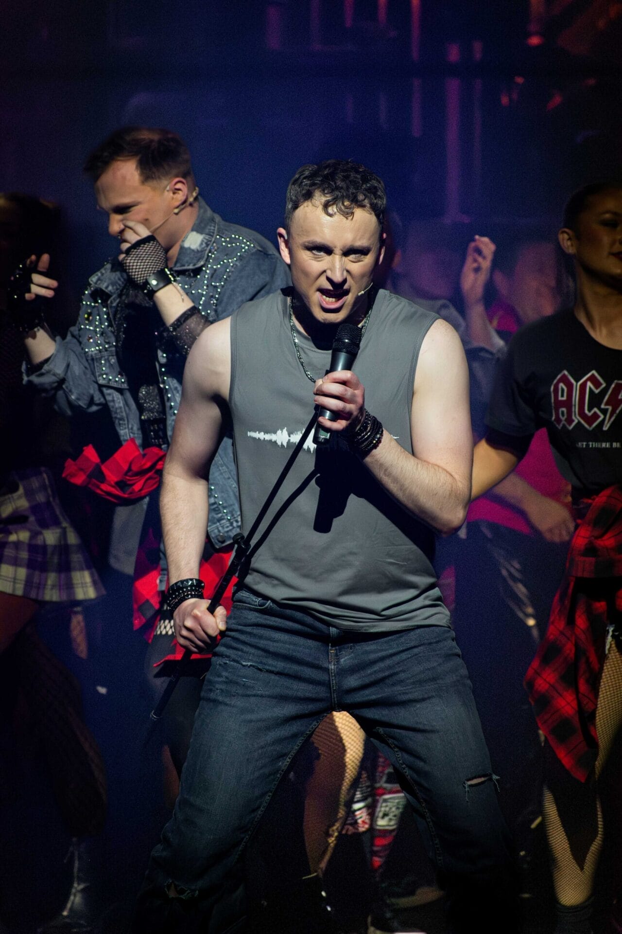 A man in a sleeveless shirt and ripped jeans sings "We Will Rock You" into a microphone on stage, surrounded by backing performers in rock-inspired clothing under dramatic lighting.