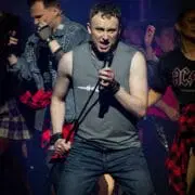 A man in a sleeveless shirt and ripped jeans sings "We Will Rock You" into a microphone on stage, surrounded by backing performers in rock-inspired clothing under dramatic lighting.