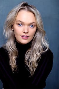 A woman with long wavy blonde hair and blue eyes is wearing a black turtleneck top, posing against a plain blue-gray background.
