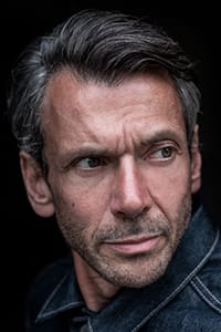A middle-aged man with short, graying hair and stubble looks to his left with a serious expression, wearing a dark denim jacket against a dark background.