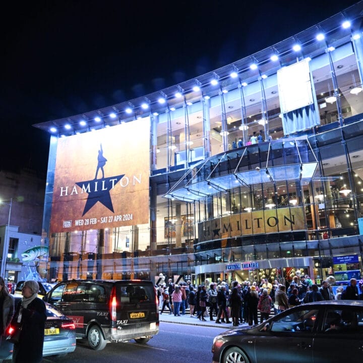 A large crowd gathers outside a brightly lit theater at night with a "Hamilton" musical banner displayed on the glass facade. Cars are parked along the street.