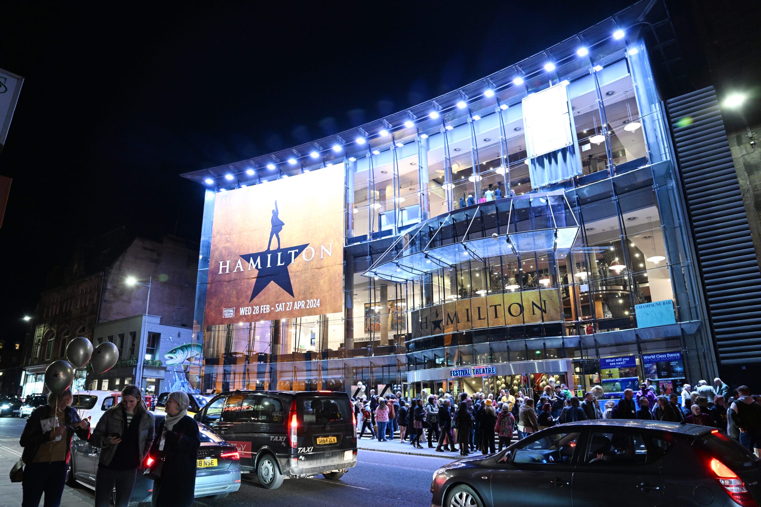 A large crowd gathers outside a brightly lit theater at night with a "Hamilton" musical banner displayed on the glass facade. Cars are parked along the street.