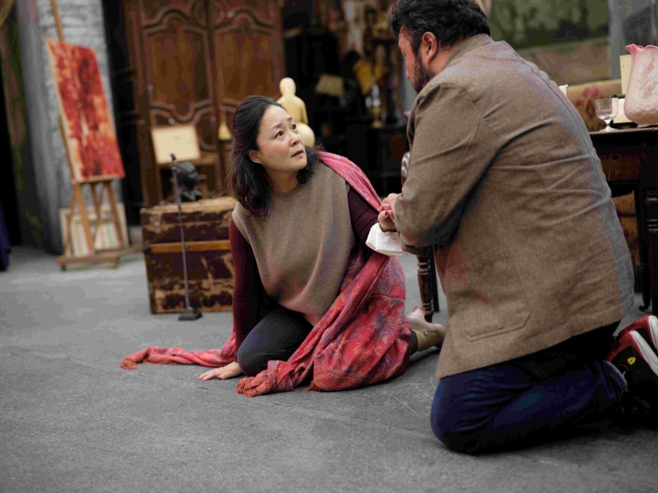A woman sits on the floor draped in a red shawl, looking up at a man kneeling in front of her in a room filled with various antique objects.