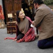 A woman sits on the floor draped in a red shawl, looking up at a man kneeling in front of her in a room filled with various antique objects.