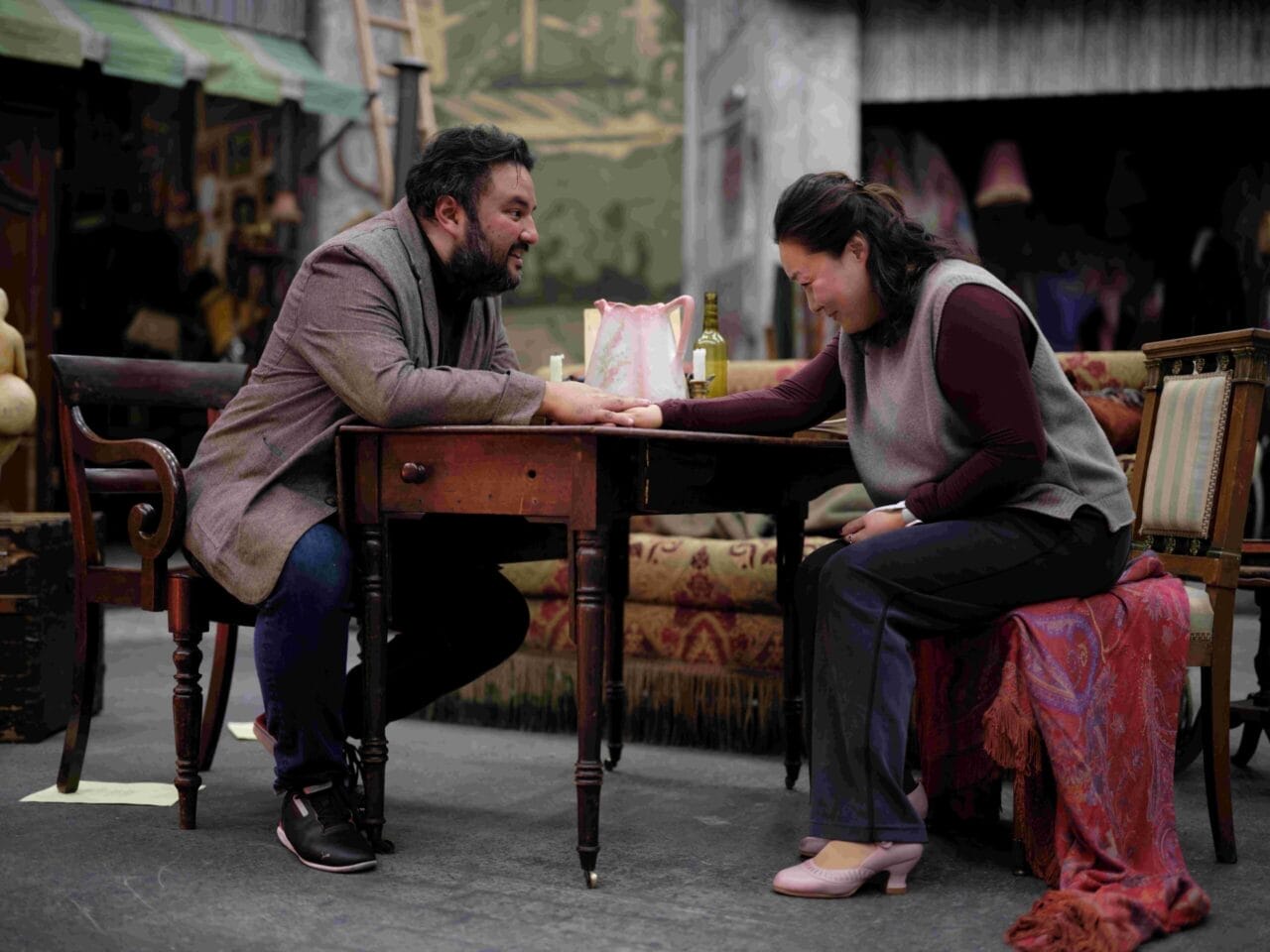 A man and a woman sit across from each other at a wooden table, holding hands, in a room decorated with vintage furniture and various objects.