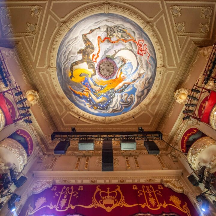 Ornate theater ceiling with a circular mural featuring abstract art, surrounded by decorated balconies with red seats and gold details.