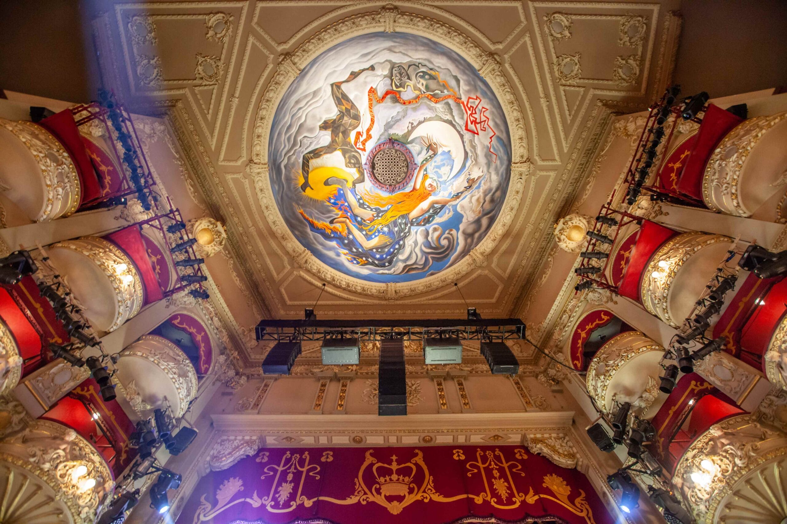 Ornate theater ceiling with a circular mural featuring abstract art, surrounded by decorated balconies with red seats and gold details.