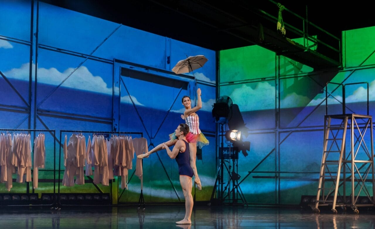 Two ballet dancers perform on stage; one lifts the other who holds an umbrella. Coats hang on racks in the background, with a painted sky and landscape set.