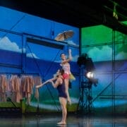 Two ballet dancers perform on stage; one lifts the other who holds an umbrella. Coats hang on racks in the background, with a painted sky and landscape set.