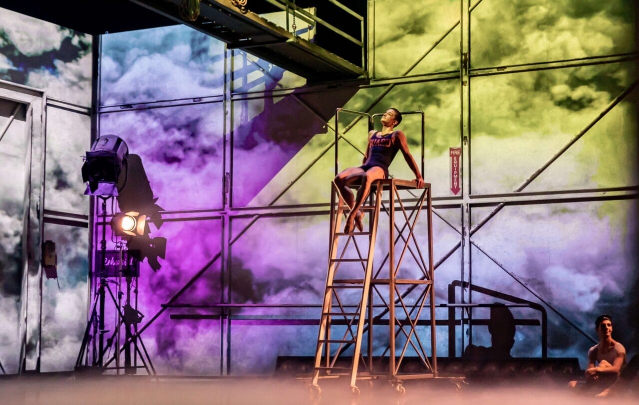 A performer in a leotard poses atop a metal ladder structure on a stage with colorful lighting and a cloudy backdrop; another person sits in the background.