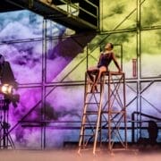 A performer in a leotard poses atop a metal ladder structure on a stage with colorful lighting and a cloudy backdrop; another person sits in the background.