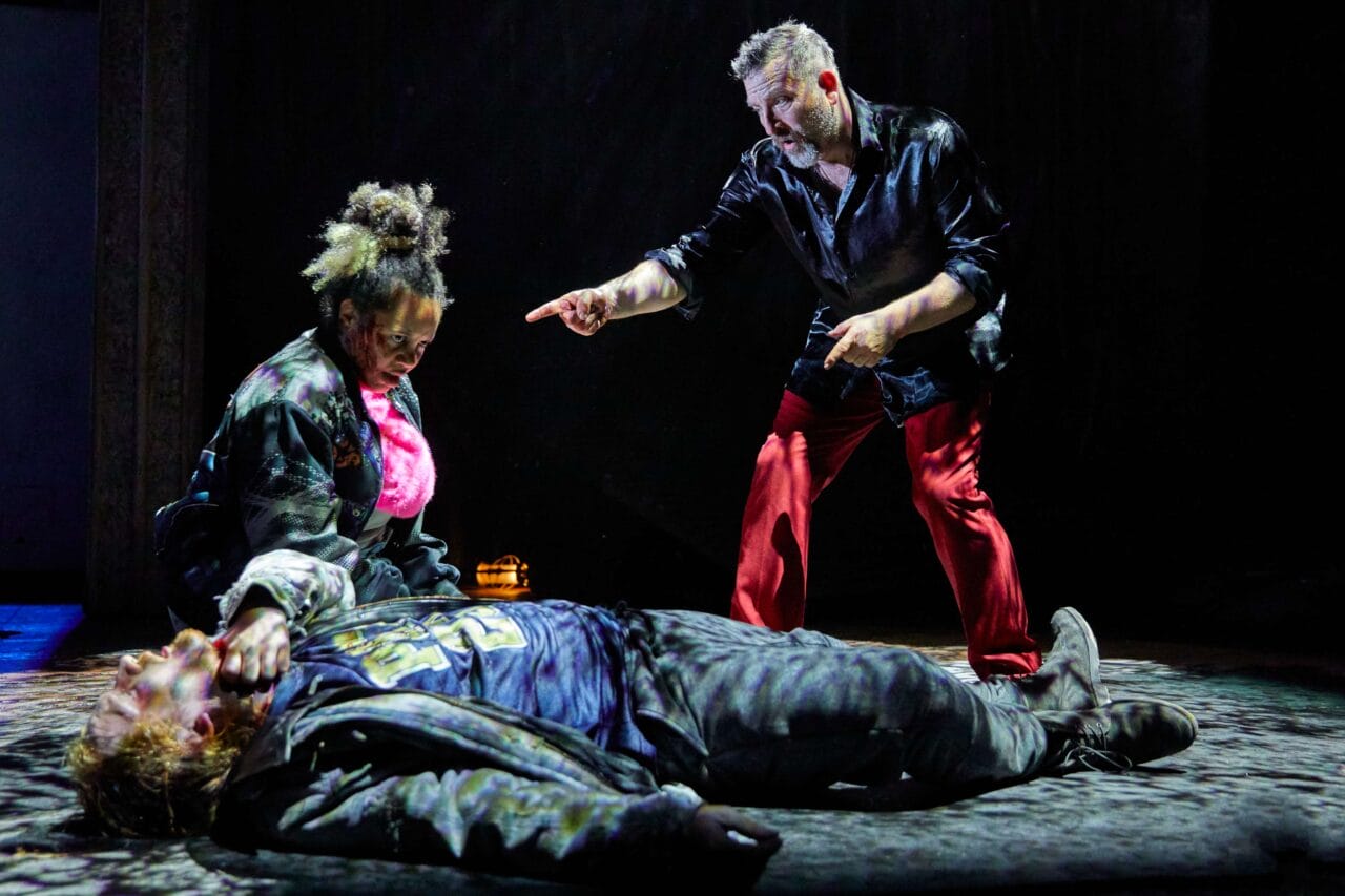 Three actors perform onstage; one person lies on the ground while another kneels beside them and a third stands, pointing, all under dramatic lighting.