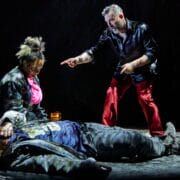 Three actors perform onstage; one person lies on the ground while another kneels beside them and a third stands, pointing, all under dramatic lighting.