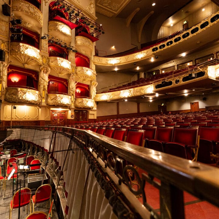 Interior of an ornate theatre with red velvet seats, gold-accented balconies, and an orchestra pit filled with music stands and chairs.