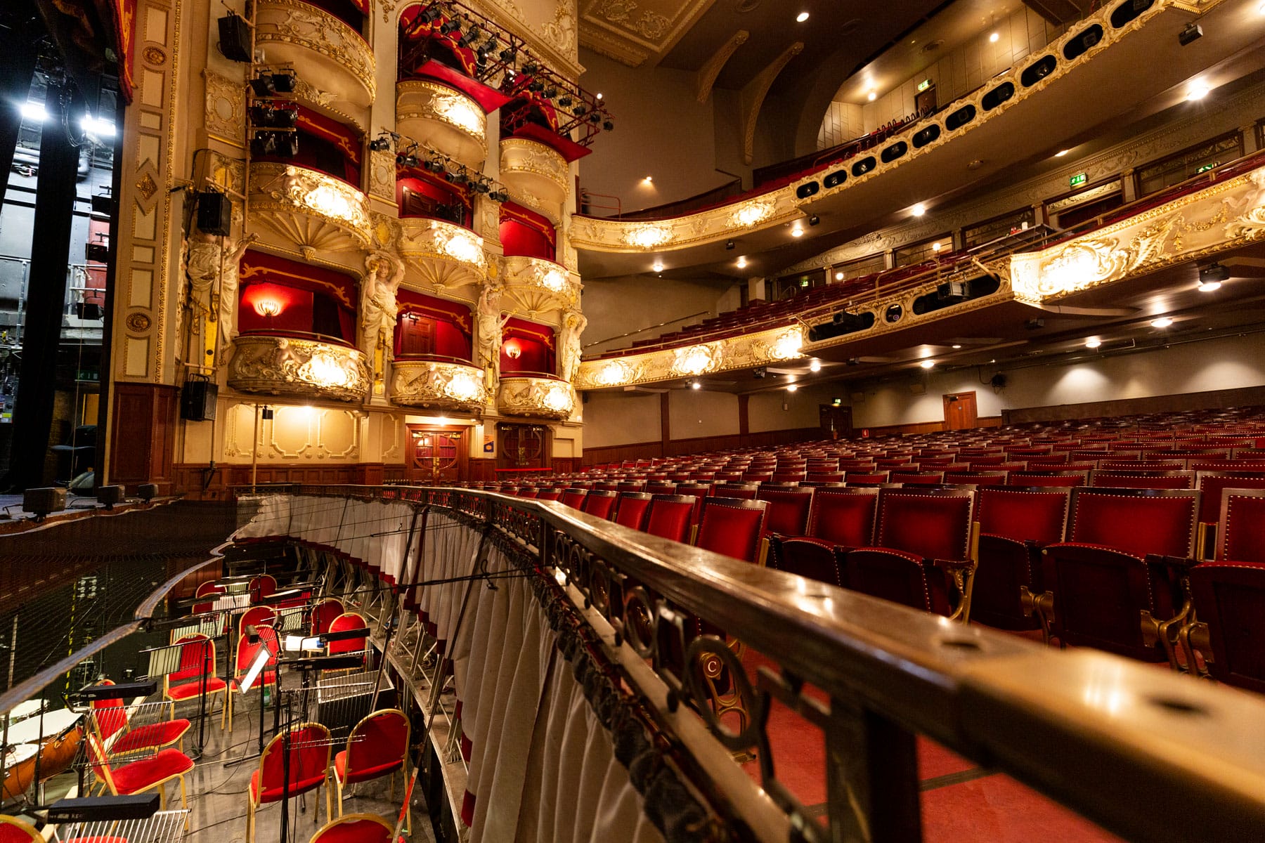 Interior of an ornate theatre with red velvet seats, gold-accented balconies, and an orchestra pit filled with music stands and chairs.