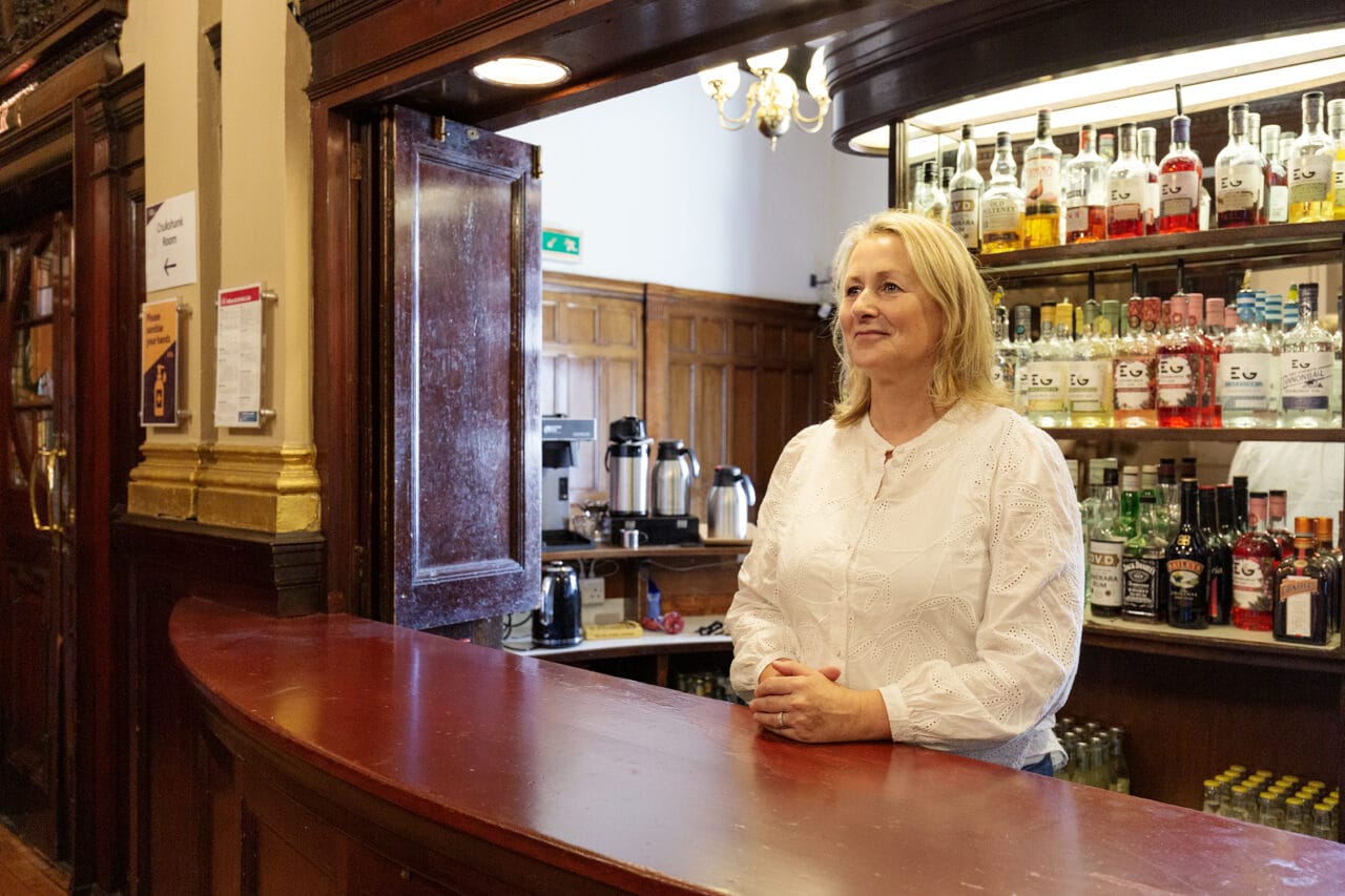A woman in a white blouse stands behind a wooden bar counter with shelves of various bottles and drinks in the background.