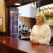 A woman in a white blouse stands behind a wooden bar counter with shelves of various bottles and drinks in the background.