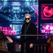 A person wearing dark sunglasses and a black coat stands in front of large, colourful digital screens. Channelling a We Will Rock You vibe, two others in bright clothing sit on either side, wires attached to their heads.