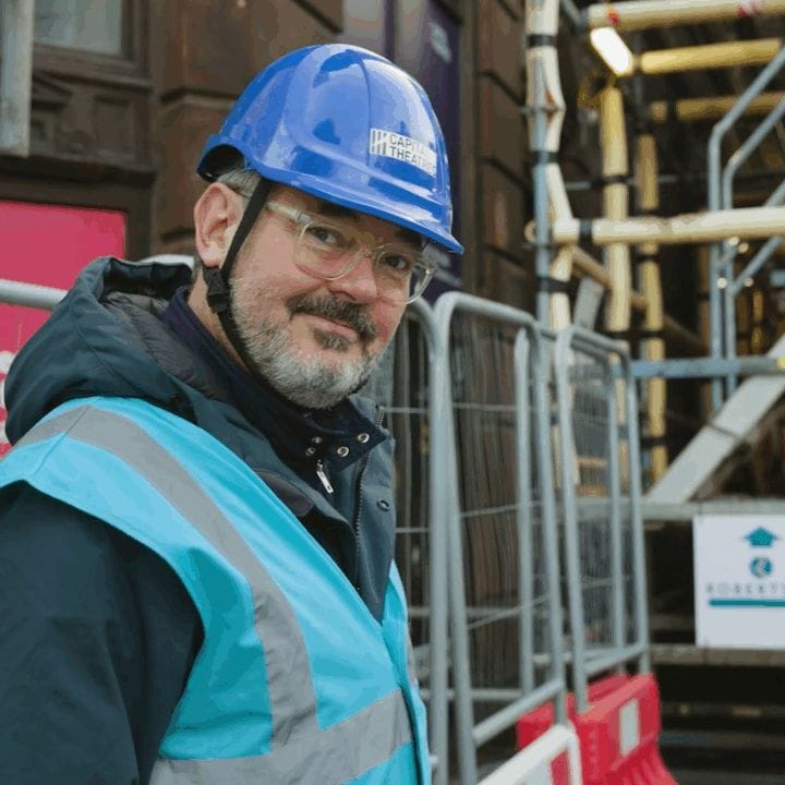 Man wearing a blue hard hat and safety vest stands near a construction site with scaffolding and pedestrian entrance signs in the background.