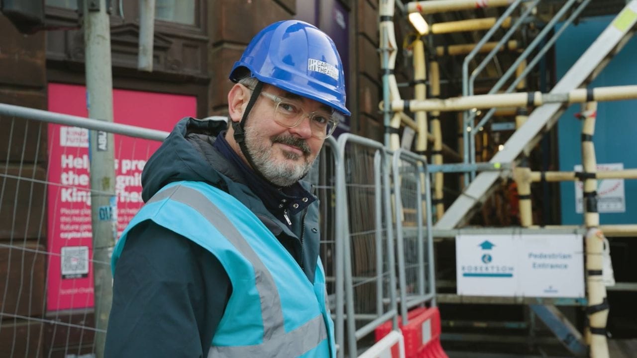 Man wearing a blue hard hat and safety vest stands near a construction site with scaffolding and pedestrian entrance signs in the background.