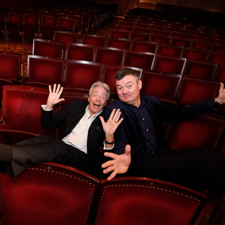 Two men in suits sit in red theatre seats with legs outstretched and arms raised, smiling energetically toward the camera in an empty auditorium.