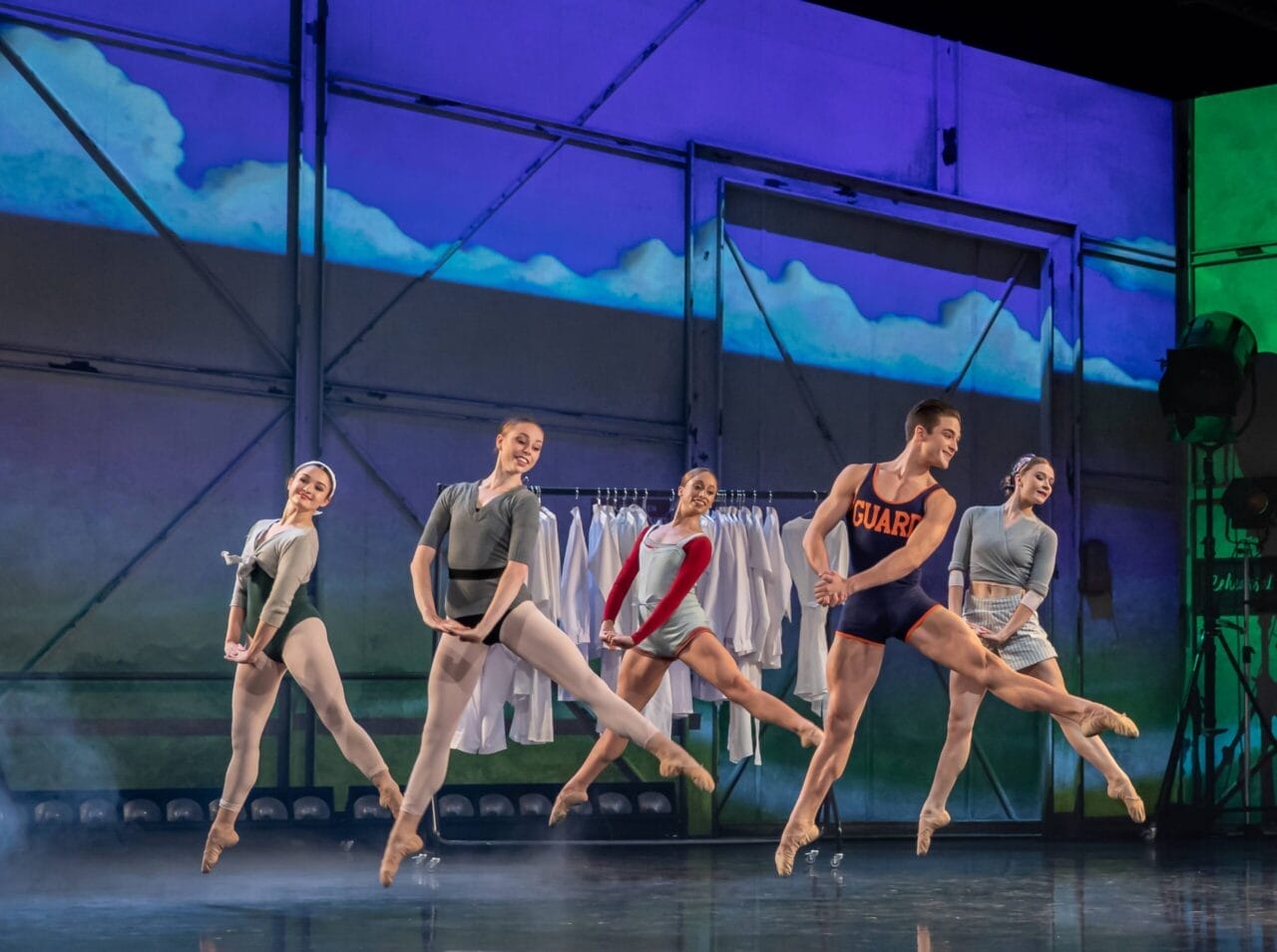 Five ballet dancers perform a synchronized leap on stage, wearing leotards and pointe shoes, with a backdrop featuring clouds and a clothing rack in the background.