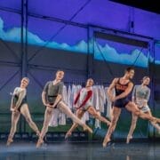 Five ballet dancers perform a synchronized leap on stage, wearing leotards and pointe shoes, with a backdrop featuring clouds and a clothing rack in the background.