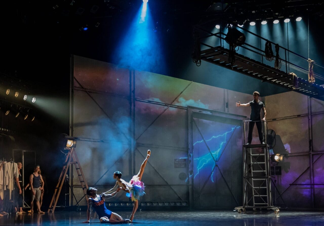 Two ballet dancers perform on stage under dramatic lighting, while two other people observe from the side and above on a metal scaffolding.