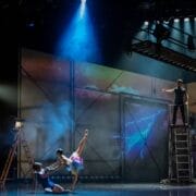 Two ballet dancers perform on stage under dramatic lighting, while two other people observe from the side and above on a metal scaffolding.