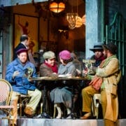 Five people in period costumes gather around a small outdoor café table, with warm lighting and a vintage setting in the background.