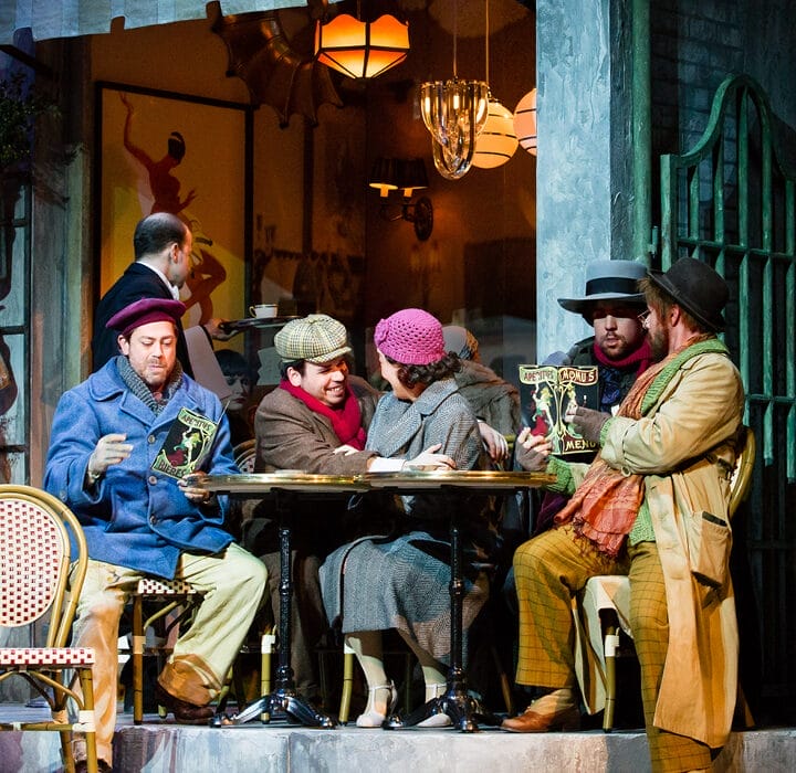 Five people in period costumes gather around a small outdoor café table, with warm lighting and a vintage setting in the background.