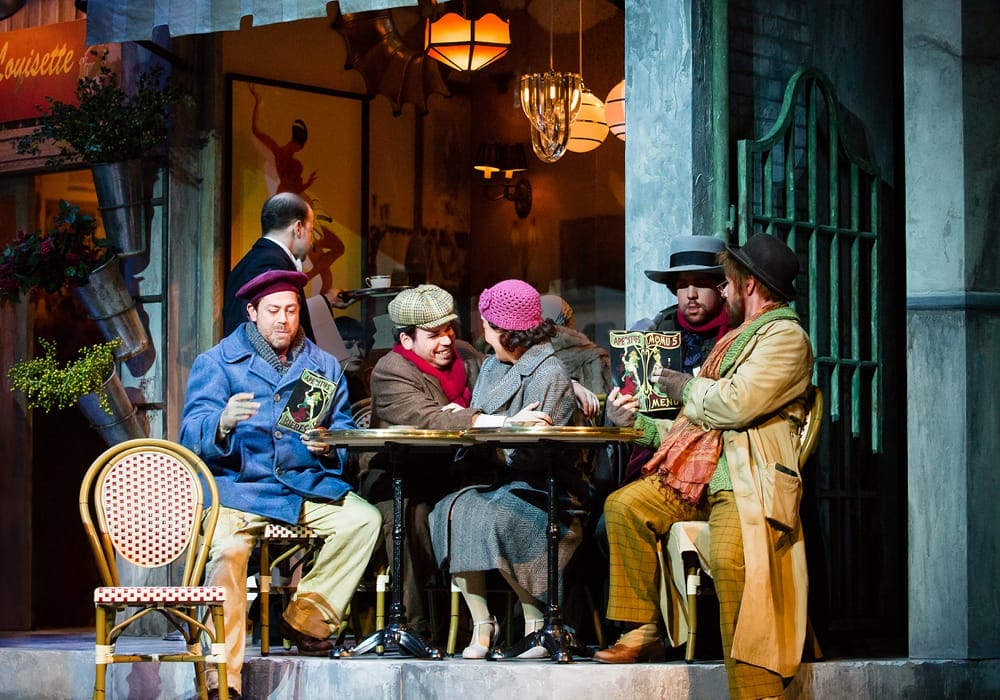 Five people in period costumes gather around a small outdoor café table, with warm lighting and a vintage setting in the background.