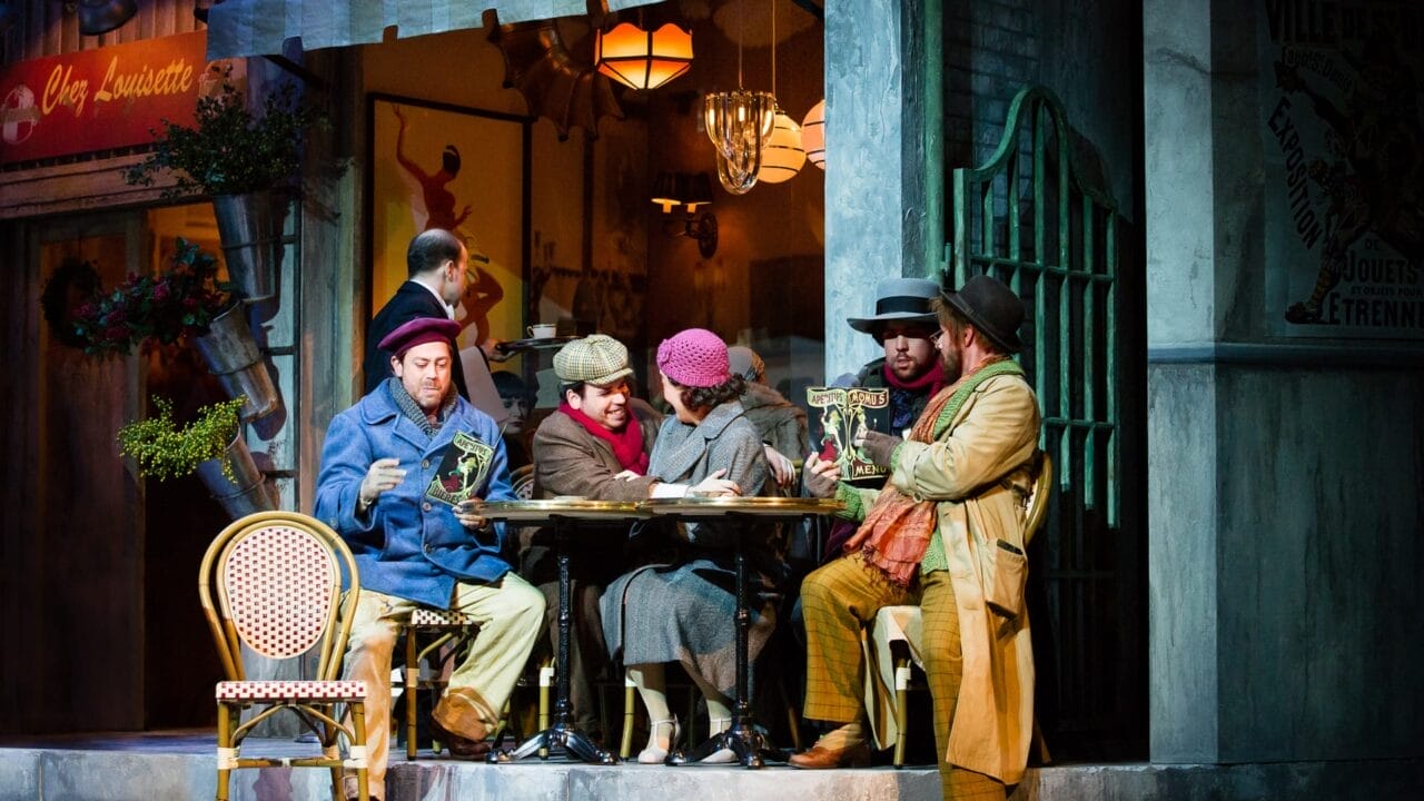 Five people in vintage attire sit at an outdoor café table, reading papers, with a waiter and another person visible inside the warmly lit restaurant.
