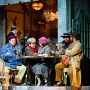 Five people in winter clothing sit around a café table outdoors, reading and conversing, with a waiter in the background and a Parisian-style café setting.