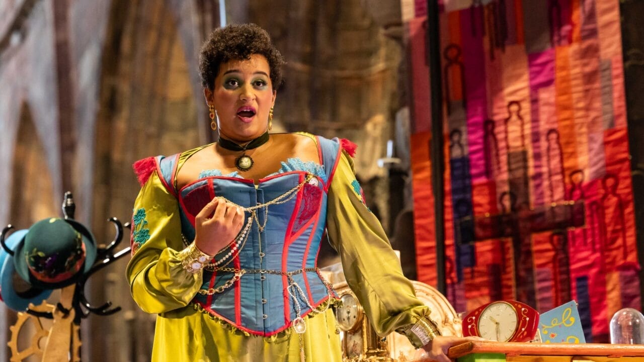 A person in a colorful, historical costume performs on stage, evoking the whimsy of L'heure espagnole, standing near a cluttered table with a clock, gears, and a vibrant fabric backdrop.