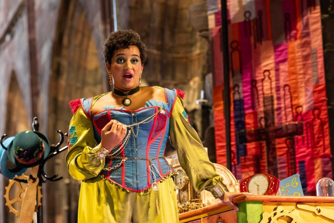 A person in a colorful, historical costume performs on stage, evoking the whimsy of L'heure espagnole, standing near a cluttered table with a clock, gears, and a vibrant fabric backdrop.