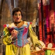 A person in a colorful, historical costume performs on stage, evoking the whimsy of L'heure espagnole, standing near a cluttered table with a clock, gears, and a vibrant fabric backdrop.
