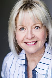 Smiling older woman with short, light blonde hair and blue eyes, wearing a white shirt with dark stripes, photographed against a neutral background.