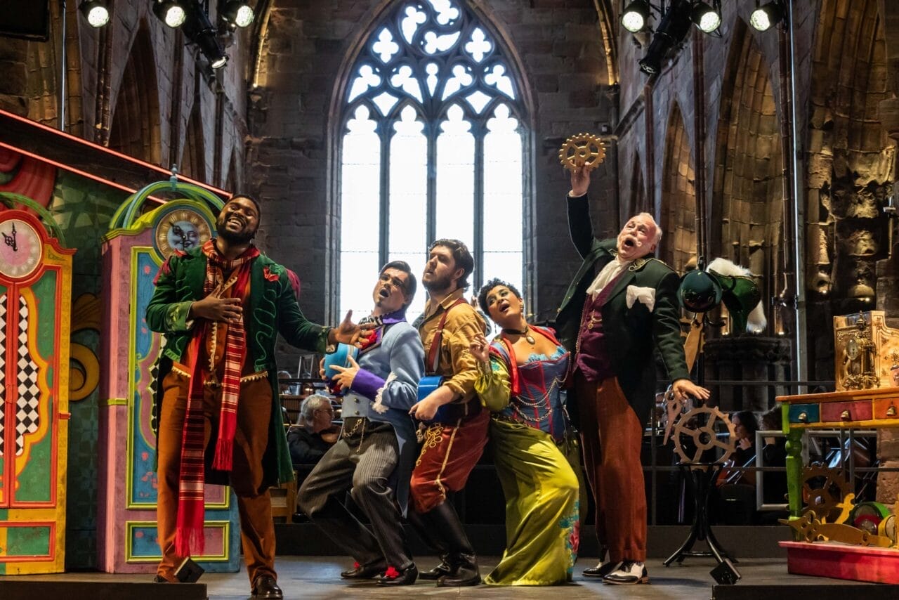 Five actors in colorful costumes perform energetically on a stage set in a grand, cathedral-like hall with arched windows, theatrical props, and echoes of The Bear and L'heure espagnole.