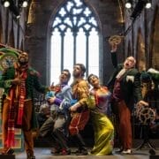 Five actors in colorful costumes perform energetically on a stage set in a grand, cathedral-like hall with arched windows, theatrical props, and echoes of The Bear and L'heure espagnole.