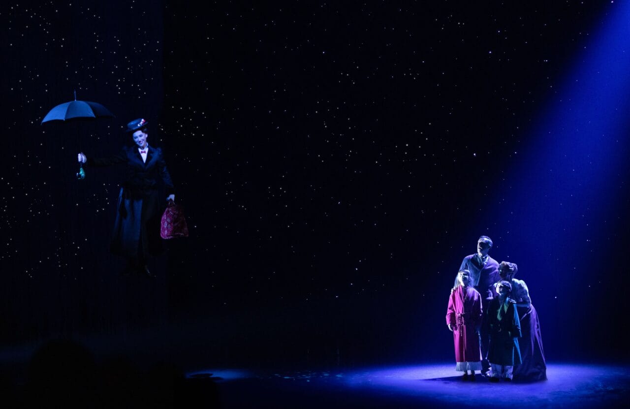 A person holding an umbrella floats in the air on a dark stage, while four people stand together in a spotlight beneath a starry backdrop.