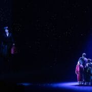 A person holding an umbrella floats in the air on a dark stage, while four people stand together in a spotlight beneath a starry backdrop.
