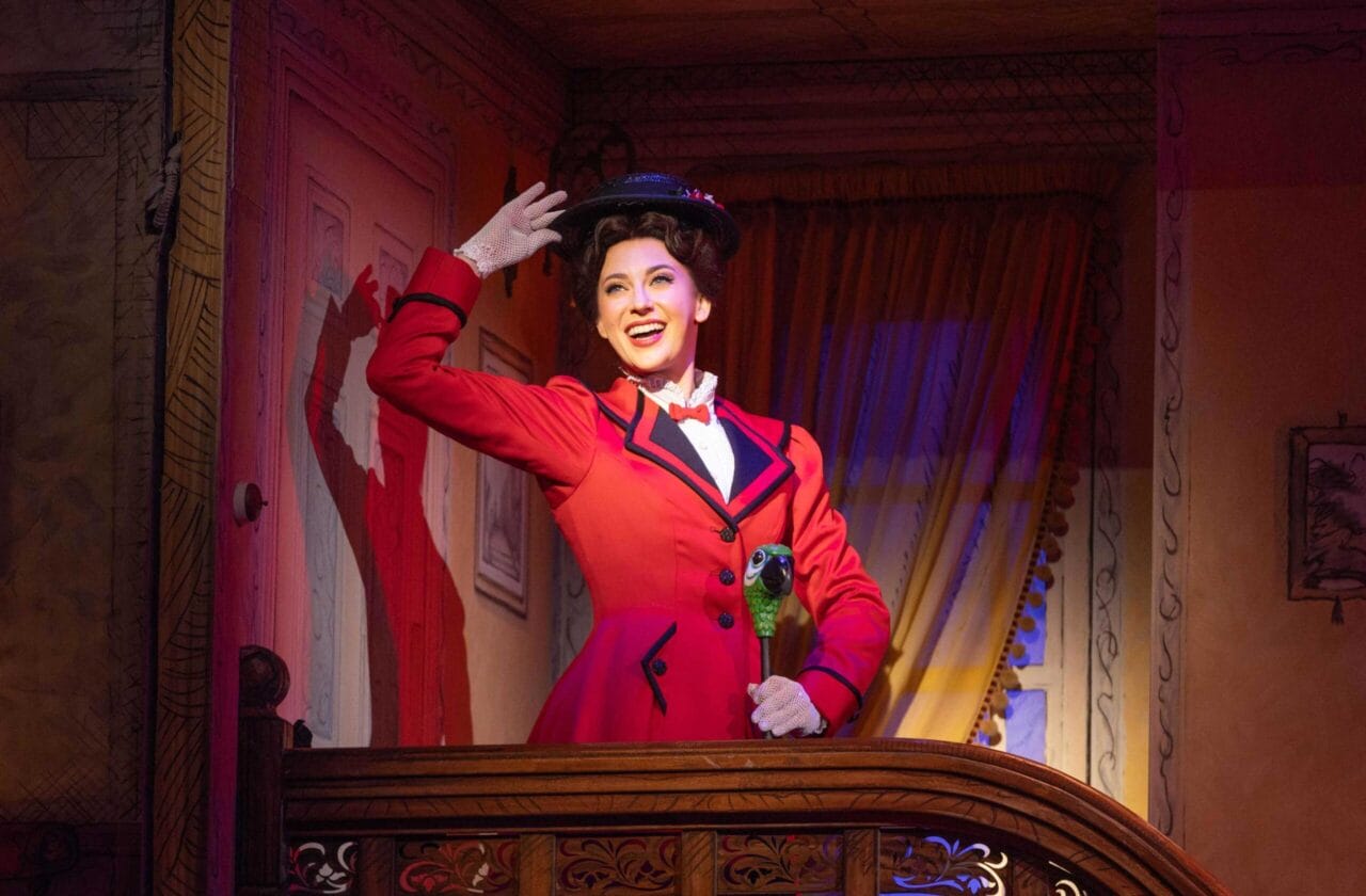 Mary Poppins in a red coat and black hat stands on a balcony indoors, smiling and raising her hand as if waving, with dramatic stage lighting highlighting her.