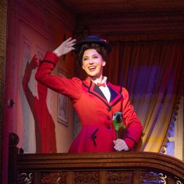 Mary Poppins in a red coat and black hat stands on a balcony indoors, smiling and raising her hand as if waving, with dramatic stage lighting highlighting her.