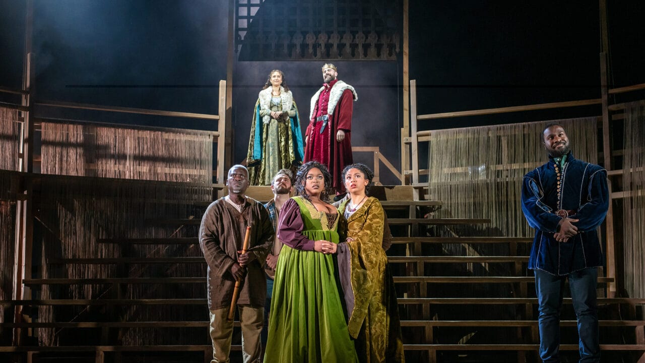 Six actors in period costumes stand on a wooden stage set with tiered steps, three on an upper level and three on the lower, under dramatic theater lighting.