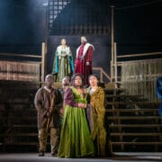 Six actors in period costumes stand on a wooden stage set with tiered steps, three on an upper level and three on the lower, under dramatic theater lighting.