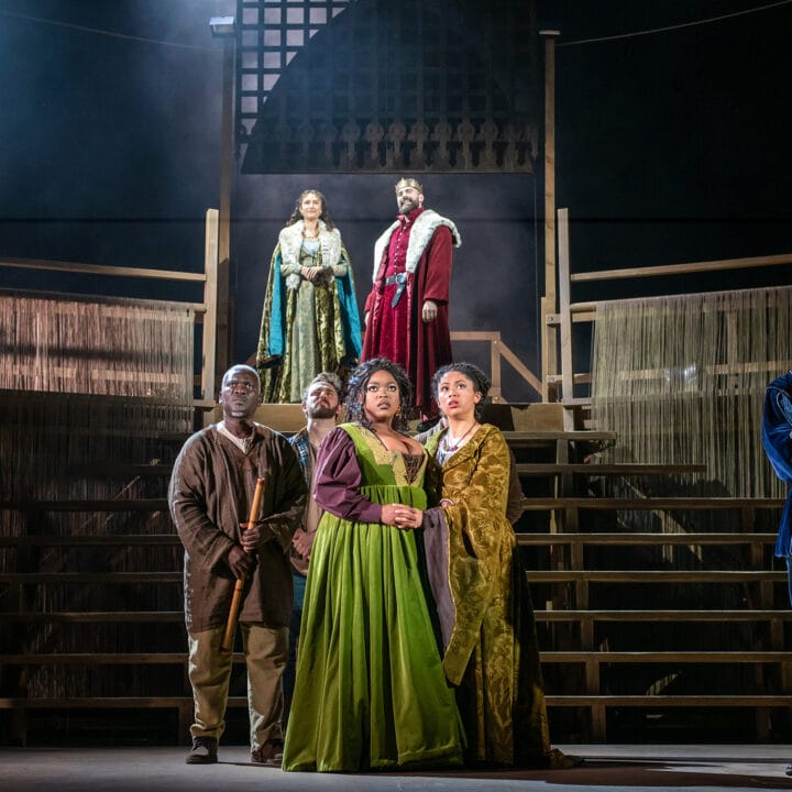 Six actors in period costumes stand on a wooden stage set with tiered steps, three on an upper level and three on the lower, under dramatic theater lighting.