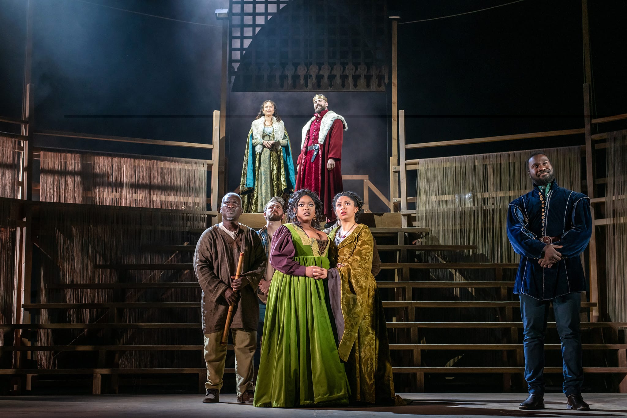 Six actors in period costumes stand on a wooden stage set with tiered steps, three on an upper level and three on the lower, under dramatic theater lighting.