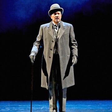 A man in a gray overcoat, bowler hat, gloves, and holding a cane stands alone on a stage with a dark blue background.